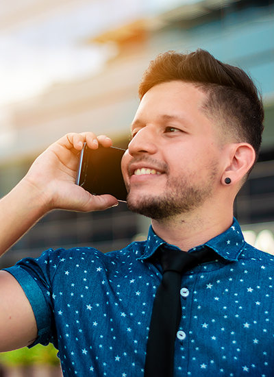 young, attractive, Hispanic Latin boy, with beard and short hair, dressed in a blue shirt and black tie, is happy talking on the phone in a business environment, with buildings in the background.