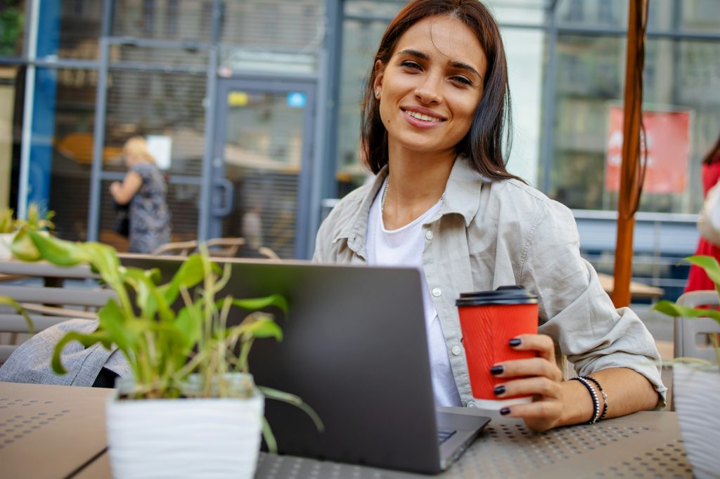 Happy beautiful woman with laptop near hi-tech business building. She drinks coffee in to go cup and