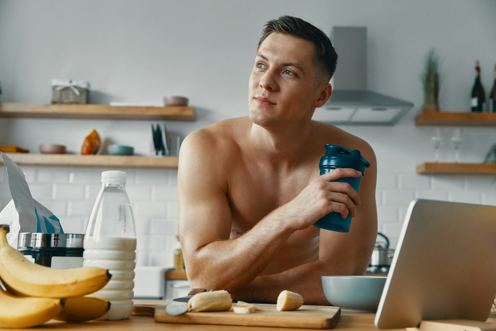 Thoughtful young man enjoying protein drink while standing at the domestic kitchen