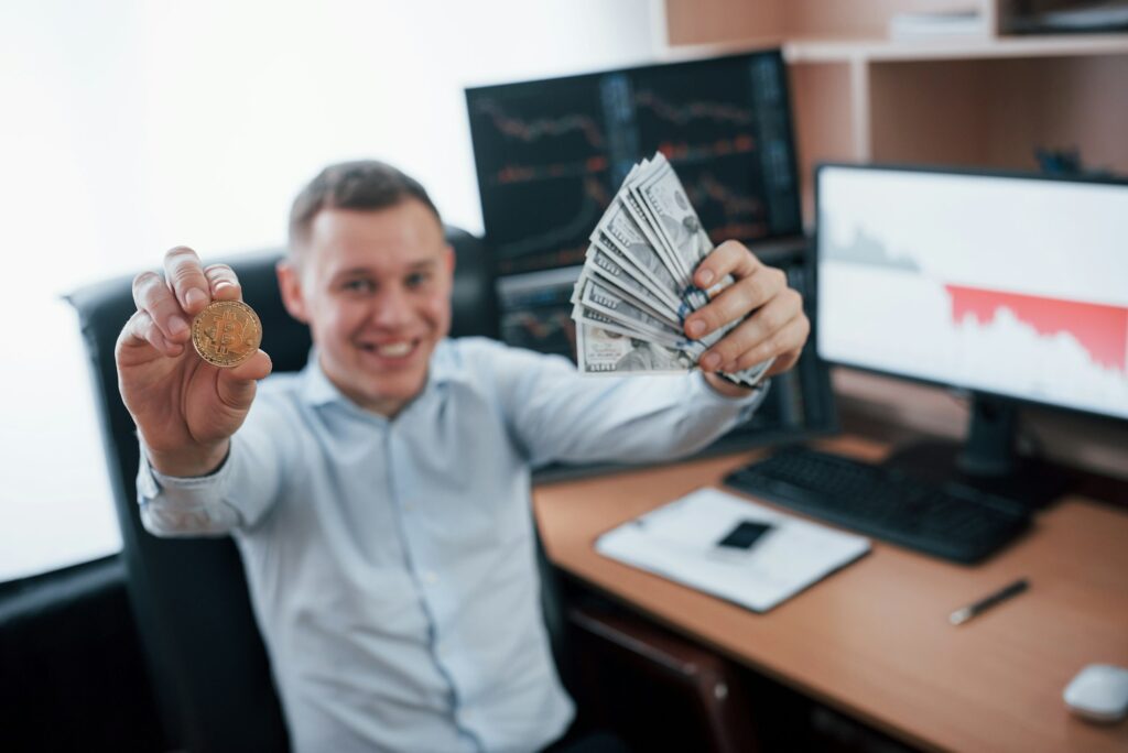 Businessman holding bitcoin and money in hands while sitting in modern office with many monitors