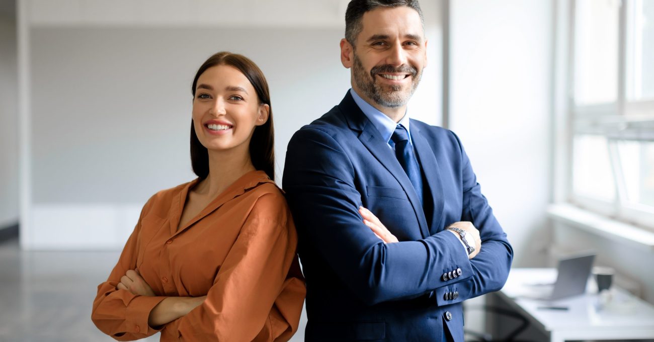 Successful happy businessman and businesswoman standing back to back with arms crossed and smiling at camera, office interior. Partnership concept